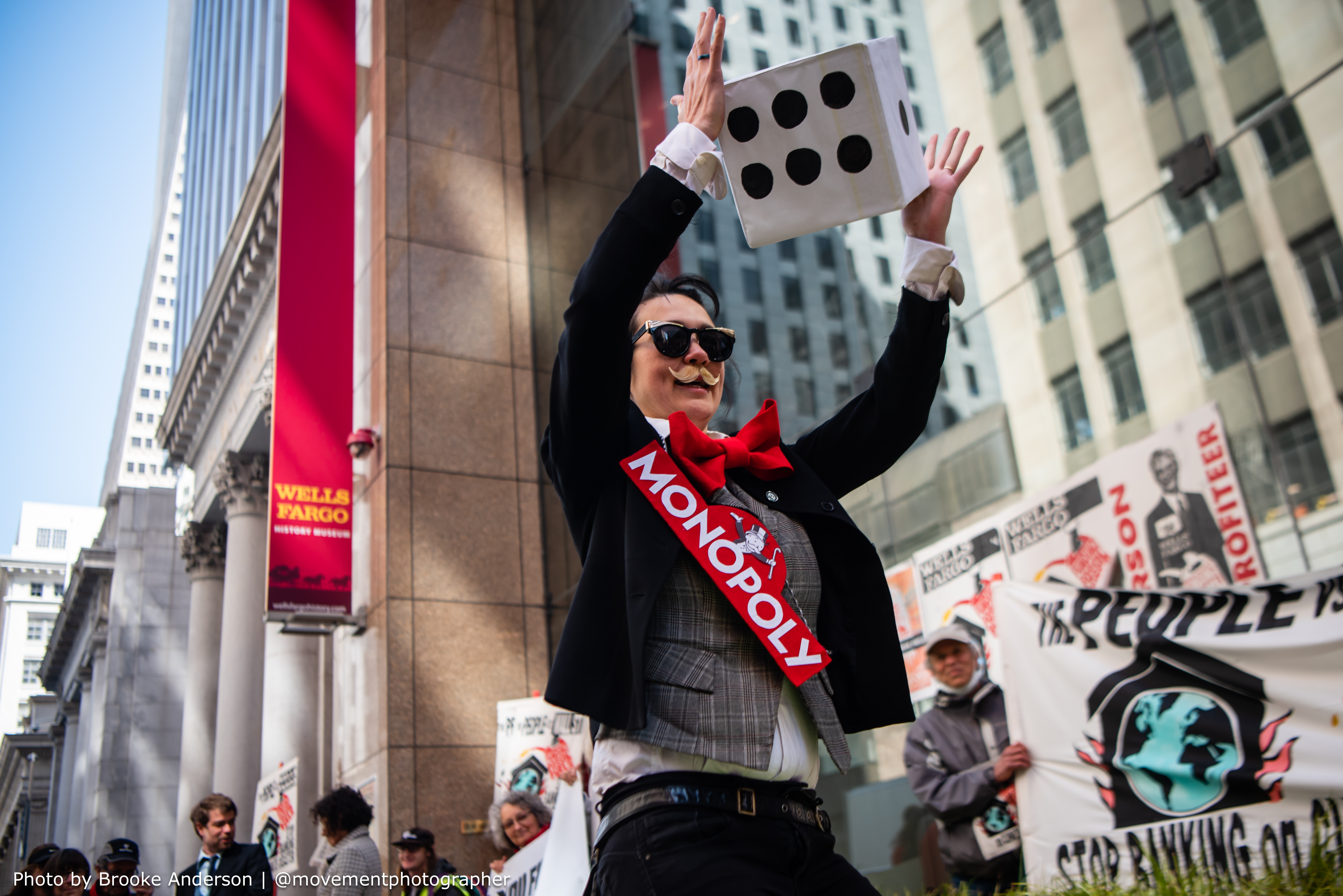 Monopoly Man Character dressed in mustache and "Monopoly" sash holds a large cartoon die in front of Wells Fargo